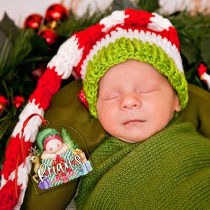 May include: A sleeping infant wearing a long, crocheted hat with red, white, and green stripes. Beside the infant is a clear acrylic ornament featuring a cartoon elf illustration and the text "Brian's First Christmas".