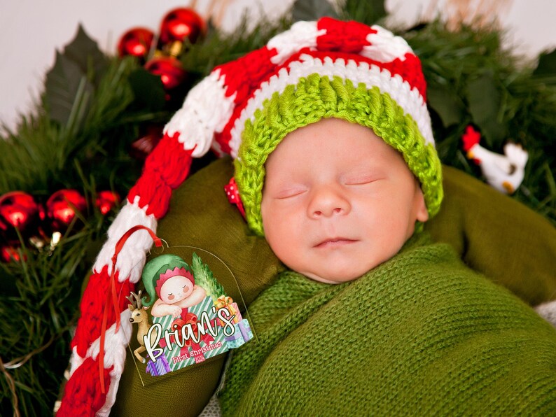 May include: A sleeping infant wearing a long, crocheted hat with red, white, and green stripes. Beside the infant is a clear acrylic ornament featuring a cartoon elf illustration and the text "Brian's First Christmas".