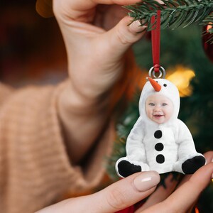 Pu&ograve; includere: Un ornamento a forma di pupazzo di neve bianco con il viso di un bambino al centro. L'ornamento ha bottoni neri e un naso di carota. L'ornamento &egrave; appeso a un nastro rosso.