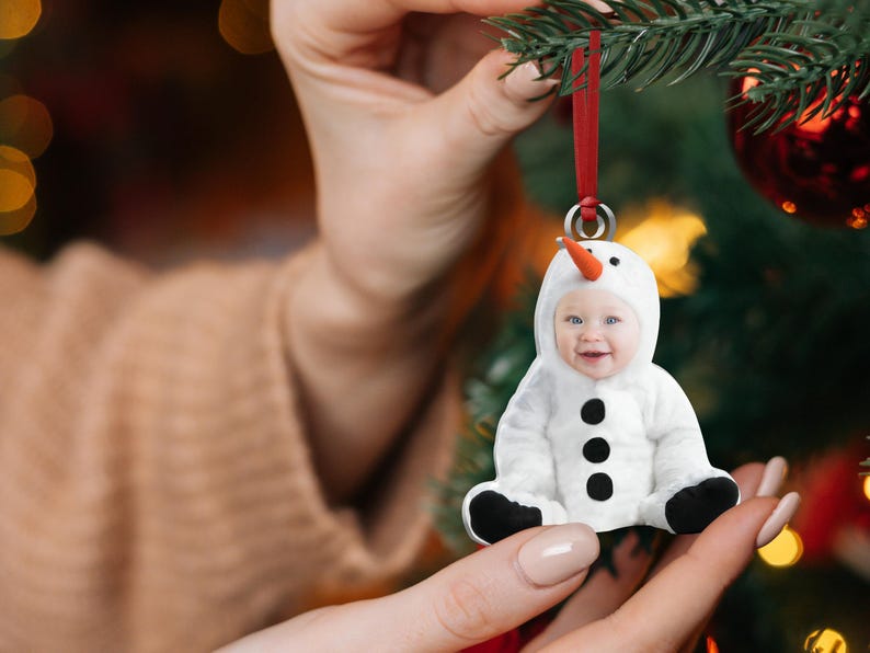 Pu&ograve; includere: Un ornamento a forma di pupazzo di neve bianco con il viso di un bambino al centro. L'ornamento ha bottoni neri e un naso di carota. L'ornamento &egrave; appeso a un nastro rosso.