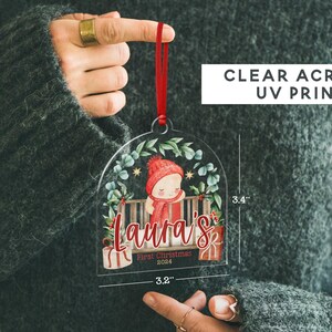May include: A clear acrylic arch-shaped Christmas ornament featuring a red ribbon, a graphic of a child in a red hat and scarf, and the text "Laura's First Christmas 2024". It measures 3.4 inches in height and 3.2 inches in width.
