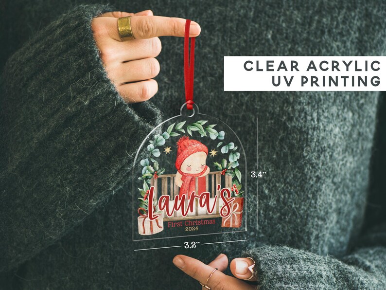 May include: A clear acrylic arch-shaped Christmas ornament featuring a red ribbon, a graphic of a child in a red hat and scarf, and the text "Laura's First Christmas 2024". It measures 3.4 inches in height and 3.2 inches in width.