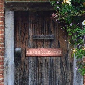 May include: A wooden door with a sign that reads "Elizabeth's Secret Place" on a rustic wooden door with a brick wall and green vines.