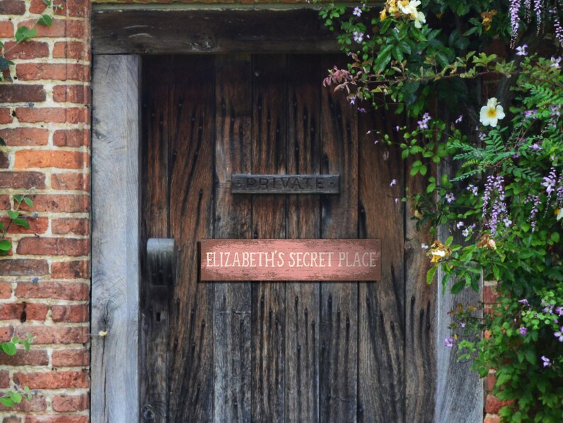 May include: A wooden door with a sign that reads "Elizabeth's Secret Place" on a rustic wooden door with a brick wall and green vines.