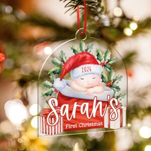 May include: A clear acrylic Christmas ornament featuring a sleeping baby in a red Santa hat with the year 2024, surrounded by holly leaves. The text reads Sarah's First Christmas, with red and white striped gift boxes at the base.