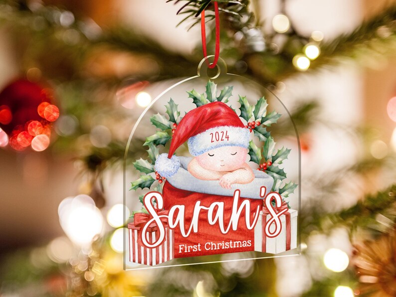 May include: A clear acrylic Christmas ornament featuring a sleeping baby in a red Santa hat with the year 2024, surrounded by holly leaves. The text reads Sarah's First Christmas, with red and white striped gift boxes at the base.
