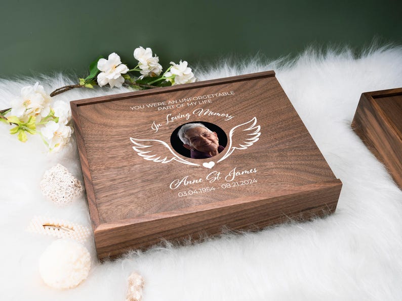 May include: A wooden box with a photo of a person and the text "YOU WERE AN UNFORGETTABLE PART OF MY LIFE In Loving Memory Anne St. James 03.04.1954-08.21.2024" engraved on the lid.
