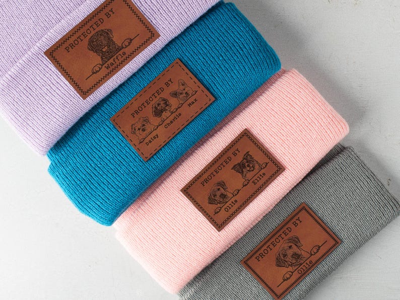 May include: Four different beanie hats with personalized leather patches. Each patch features a dog's photo and name. The hats are in various colors: purple, blue, pink, and gray.