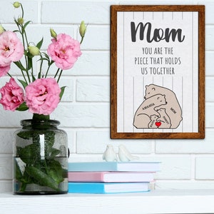May include: A framed print with a white background and brown wood frame. The print features a graphic of a family of bears hugging each other. The text "Mom You are the piece that holds us together" is written above the bears. The bears are labeled "Mom", "Amanda", "Haley", and "Robert".