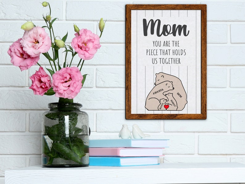 May include: A framed print with a white background and brown wood frame. The print features a graphic of a family of bears hugging each other. The text "Mom You are the piece that holds us together" is written above the bears. The bears are labeled "Mom", "Amanda", "Haley", and "Robert".