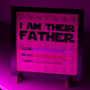 May include: A framed pink print with the text "I AM THEIR FATHER" and three lightsabers. The names Noah, Lucas, and Sophia are below the lightsabers. The background has Star Wars character illustrations.