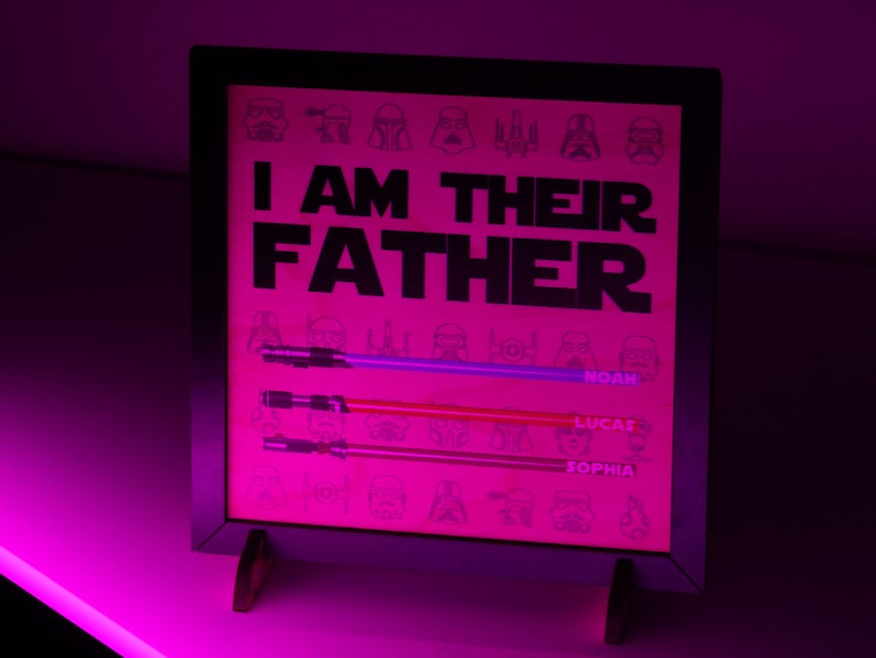 May include: A framed pink print with the text "I AM THEIR FATHER" and three lightsabers. The names Noah, Lucas, and Sophia are below the lightsabers. The background has Star Wars character illustrations.