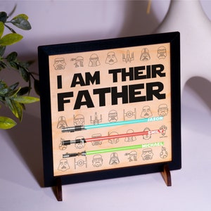 May include: Wooden framed sign with the text "I AM THEIR FATHER" and Star Wars character illustrations. The sign features three light sabers in blue, red, and green, with names below. The sign is displayed on a white surface.