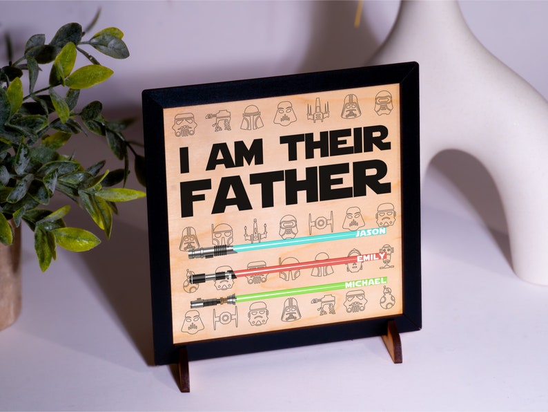 May include: Wooden framed sign with the text "I AM THEIR FATHER" and Star Wars character illustrations. The sign features three light sabers in blue, red, and green, with names below. The sign is displayed on a white surface.