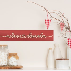 May include: Red wooden sign with the names "Melanie" and "Alexander" written in cursive with a heart symbol between the names. The sign is hanging on a white wall with a branch with red and white gingham heart ornaments hanging from it.