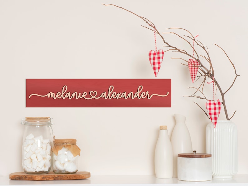 May include: Red wooden sign with the names "Melanie" and "Alexander" written in cursive with a heart symbol between the names. The sign is hanging on a white wall with a branch with red and white gingham heart ornaments hanging from it.