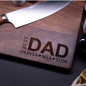 May include: A wooden cutting board with the words "Best Dad" engraved on it. The names Olivia, Mia, and Tom are also engraved on the board, along with heart symbols.