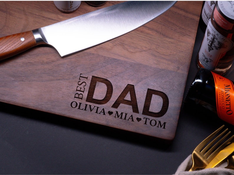 May include: A wooden cutting board with the words "Best Dad" engraved on it. The names Olivia, Mia, and Tom are also engraved on the board, along with heart symbols.