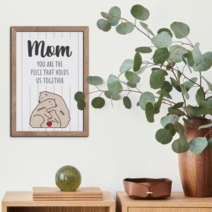 May include: A framed print with a white background and black text that reads "Mom you are the piece that holds us together."  The print features a cartoon illustration of three brown bears hugging with the names "Sophia", "Alice", and "Mia" written on them.