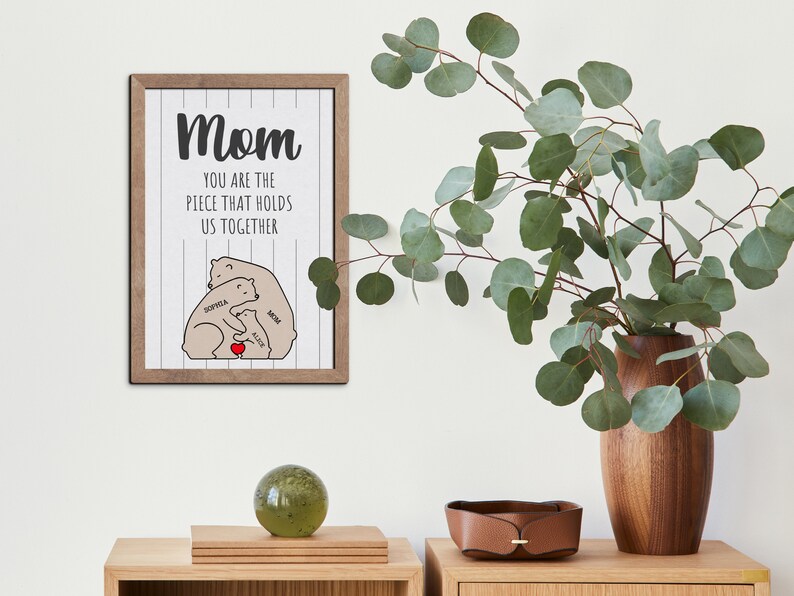 May include: A framed print with a white background and black text that reads "Mom you are the piece that holds us together."  The print features a cartoon illustration of three brown bears hugging with the names "Sophia", "Alice", and "Mia" written on them.