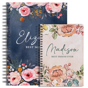 May include: Two spiral bound notebooks with floral designs. The top notebook is navy blue with the text "Eliza Best Mom" and the bottom notebook is peach with the text "Madison Best Friend Ever".