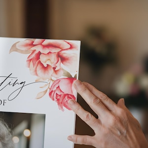 May include: A white framed print with the words "Celebrating the Life of" in elegant script. The upper right corner features watercolor-style pink and red floral artwork. A hand gently touches the frame.