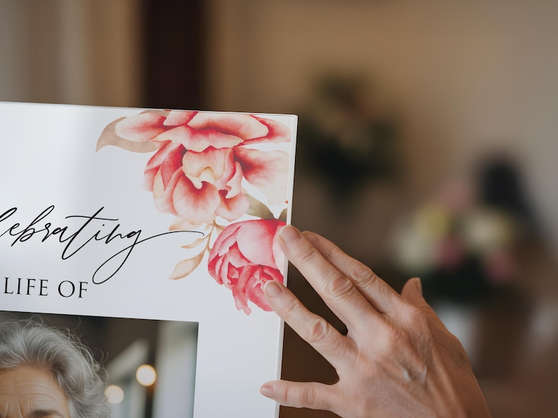 May include: A white framed print with the words "Celebrating the Life of" in elegant script. The upper right corner features watercolor-style pink and red floral artwork. A hand gently touches the frame.