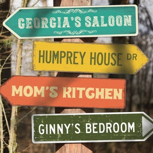 May include: Four vintage-style wooden signs with arrow shapes point in different directions. The signs are painted in various colors and have text that reads "Georgia's Saloon", "Humprey House Dr", "Mom's Kitchen", and "Ginny's Bedroom".