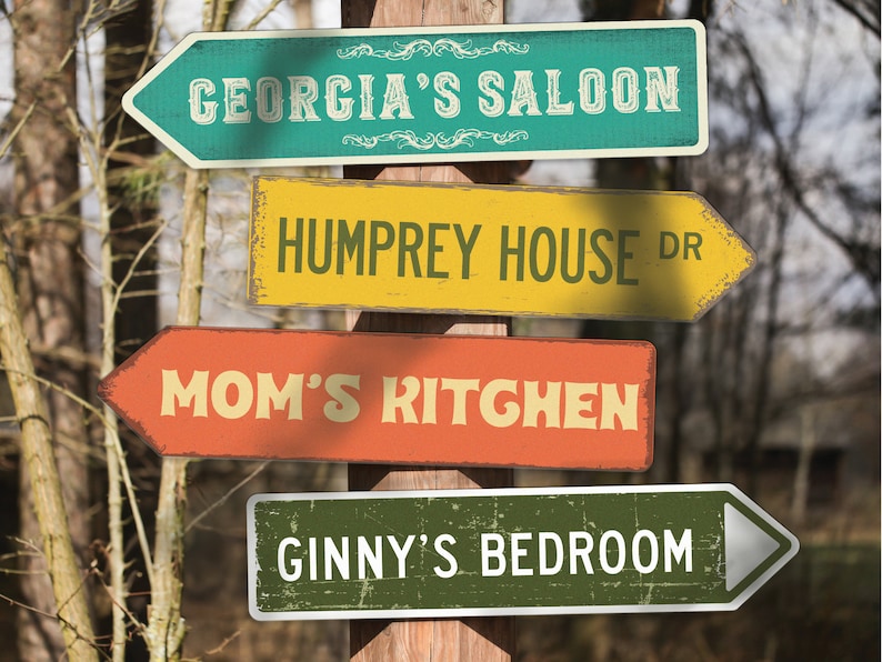 May include: Four vintage-style wooden signs with arrow shapes point in different directions. The signs are painted in various colors and have text that reads "Georgia's Saloon", "Humprey House Dr", "Mom's Kitchen", and "Ginny's Bedroom".