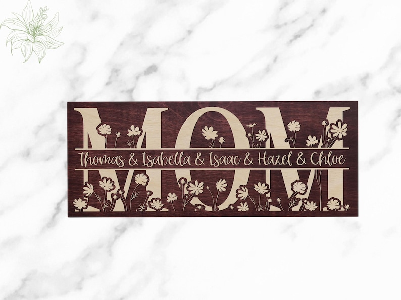May include: A wooden sign with the word "MOM" in large, beige letters on a dark brown background. The letters are surrounded by a floral design. The text "Thomas & Isabella & Isaac & Hazel & Chloe" is written in beige letters below the word "MOM".