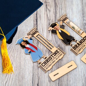Op de afbeelding: Houten afstudeercadeaus met cartoon afgestudeerden, gepersonaliseerd met namen en universiteitsdetails. De cadeaus bevatten de tekst "Gefeliciteerd! Klas van 2025". Een blauwe afstudeerhoed met een gele kwast is ook zichtbaar.