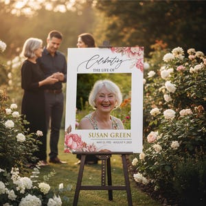 May include: A memorial sign on a wooden easel, featuring a photo of Susan Green, with the text "Celebrating the Life of Susan Green" and dates May 12, 1952 - August 9, 2023. The sign is decorated with pink and red floral accents.