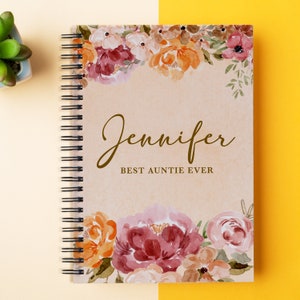 May include: A spiral bound notebook with a floral design on the cover. The cover is beige with watercolor flowers in shades of pink, orange, and brown. The text "Jennifer Best Auntie Ever" is printed in brown on the cover.