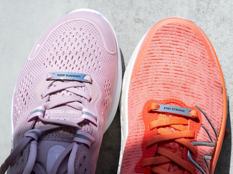Puede incluir: Dos pares de zapatillas de running rosas y naranjas con cordones de metal plateado que tienen las palabras "Keep Running!" y "Stay Strong!"
