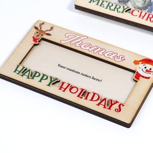 May include: A wooden Christmas photo frame with a red and green "Happy Holidays" inscription, a reindeer and snowman cutout, and a space for a personalized name. The frame says "Thomas" and has a space for a custom note.