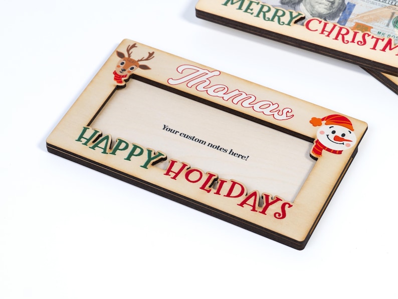 May include: A wooden Christmas photo frame with a red and green "Happy Holidays" inscription, a reindeer and snowman cutout, and a space for a personalized name. The frame says "Thomas" and has a space for a custom note.