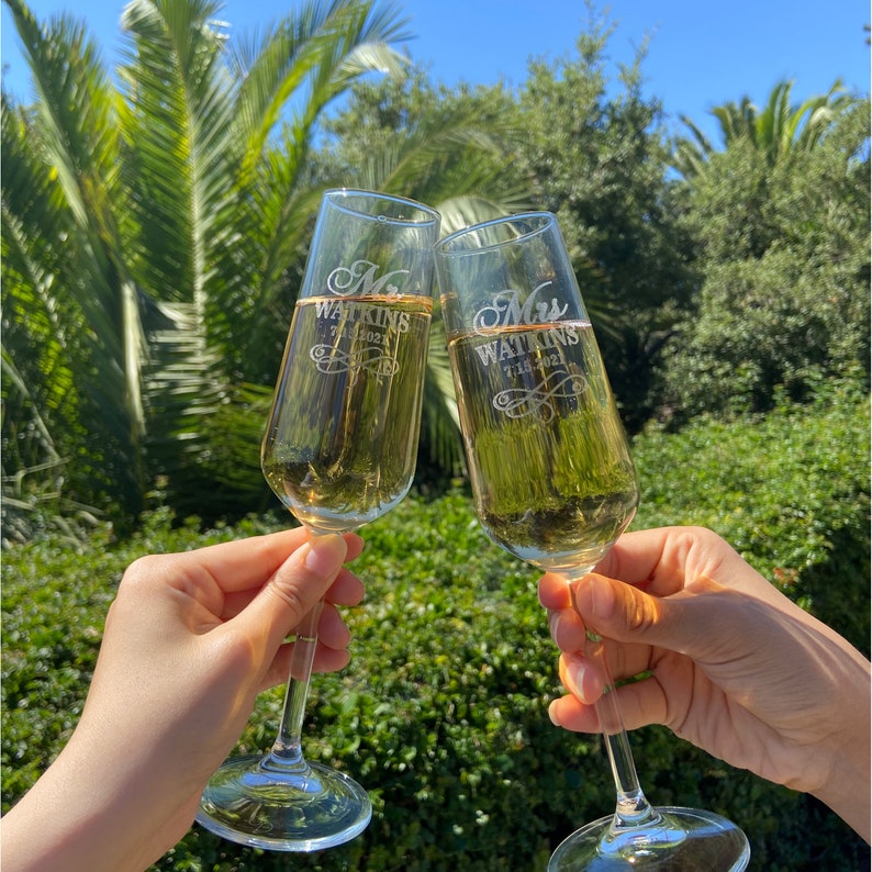 May include: Two champagne flutes with the words "Mr. Watkins 7.16.2021" and "Mrs. Watkins 7.16.2021" etched on them. The flutes are filled with champagne and are being held up in a toast.