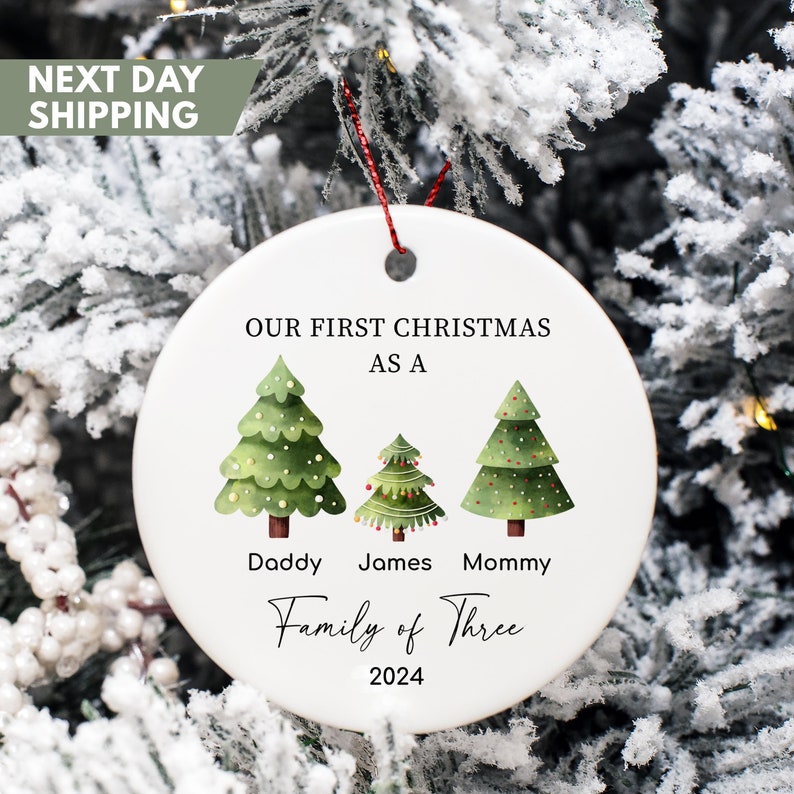 May include: White ceramic Christmas ornament with green, red, and white illustrated Christmas trees. The ornament reads "Our First Christmas as a Family of Three 2024".