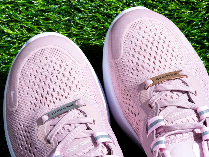 Puede incluir: Un par de zapatillas de running rosas con cordones blancos y un charm de cord&oacute;n de metal plateado y dorado. Los charms tienen el texto "Just Keep Running" y "Keep Going Mom!"