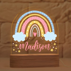May include: A rainbow-shaped night light with the name "Madison" in pink script. The rainbow features pastel colors and is set on a wooden base. The light is illuminated, creating a warm glow. Stars and clouds are also included in the design.