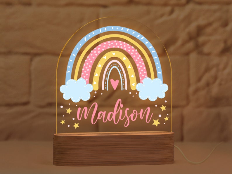 May include: A rainbow-shaped night light with the name "Madison" in pink script. The rainbow features pastel colors and is set on a wooden base. The light is illuminated, creating a warm glow. Stars and clouds are also included in the design.