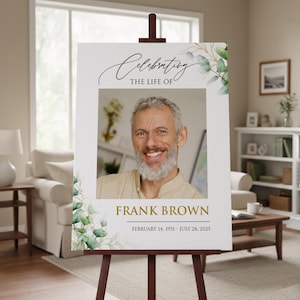May include: A memorial sign on a wooden easel. The sign features a photo of a smiling man, with the text "Celebrating the Life of Frank Brown" and his birth and passing dates. Eucalyptus leaves and branches decorate the edges.