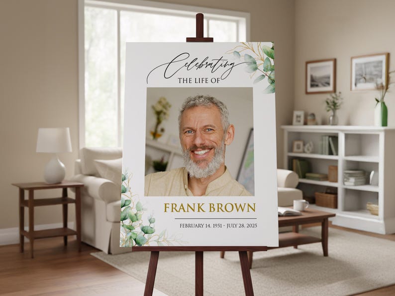 May include: A memorial sign on a wooden easel. The sign features a photo of a smiling man, with the text "Celebrating the Life of Frank Brown" and his birth and passing dates. Eucalyptus leaves and branches decorate the edges.