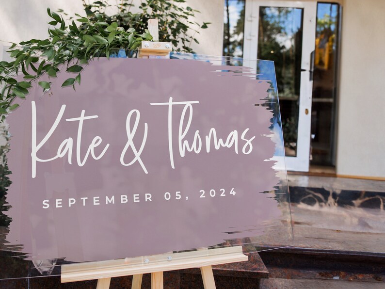 Pu&ograve; includere: Un cartello nuziale trasparente su un cavalletto di legno. Il cartello presenta i nomi "Kate & Thomas" in corsivo bianco su un design a pennellate color malva. La data "SEPTEMBER 05, 2024" &egrave; stampata sotto. Sullo sfondo si vede del verde.