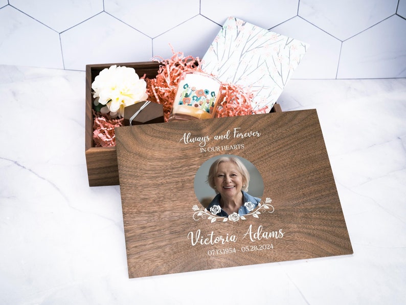May include: A wooden box with a lid that opens to reveal a photo of a woman, a candle, and tissue paper. The lid is engraved with the text "Always and Forever in Our Hearts" and "Victoria Adams 07.13.1954 - 05.28.2024".