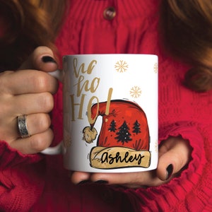 Puede incluir: Taza de cer&aacute;mica blanca con una ilustraci&oacute;n de gorro de Pap&aacute; Noel rojo y el texto "Ho Ho Ho!" en letras doradas. El nombre "Ashley" est&aacute; escrito en letras doradas en una pancarta debajo del gorro.