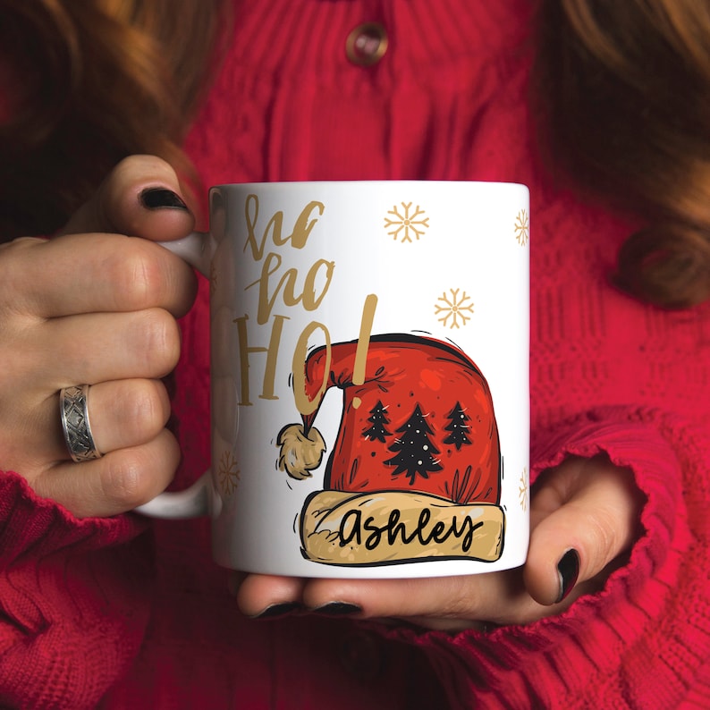 Puede incluir: Taza de cer&aacute;mica blanca con una ilustraci&oacute;n de gorro de Pap&aacute; Noel rojo y el texto "Ho Ho Ho!" en letras doradas. El nombre "Ashley" est&aacute; escrito en letras doradas en una pancarta debajo del gorro.