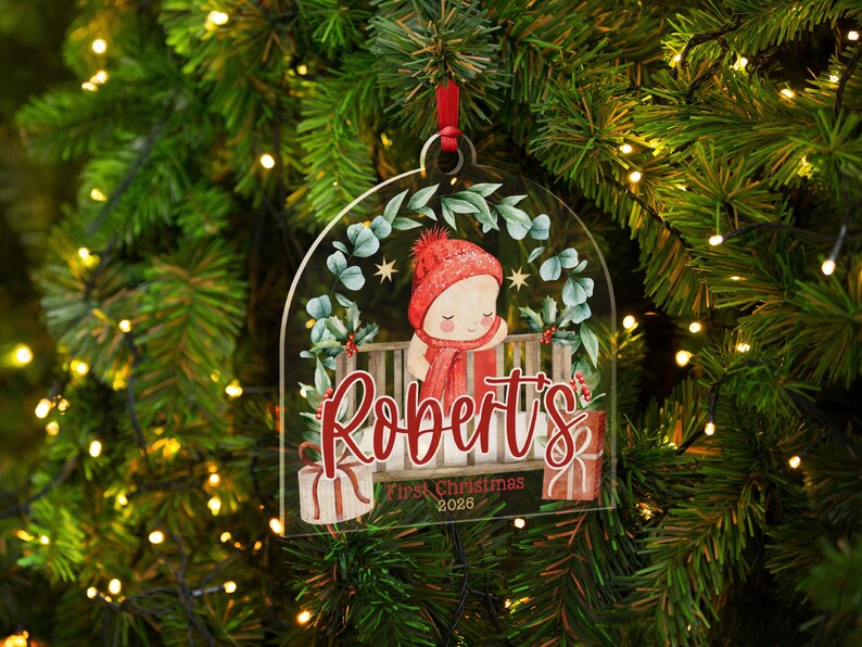 May include: A clear, arch-shaped acrylic ornament featuring a printed illustration of a baby in a red knit hat and scarf inside a crib. The text reads Robert's First Christmas 2026, surrounded by a green eucalyptus wreath and red gift boxes.