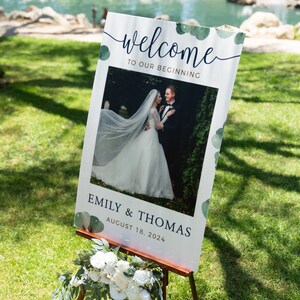 May include: A wedding welcome sign on a wooden easel. The sign features the words "welcome to our beginning" in elegant script, with a photo of a bride and groom. The sign also includes the names "Emily & Thomas" and the date "August 18, 2024".