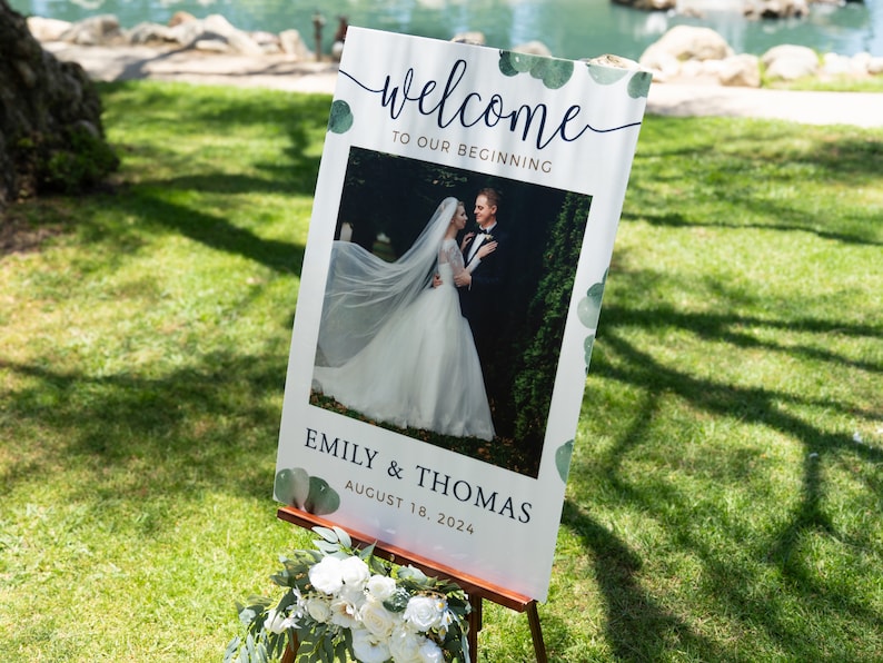 May include: A wedding welcome sign on a wooden easel. The sign features the words "welcome to our beginning" in elegant script, with a photo of a bride and groom. The sign also includes the names "Emily & Thomas" and the date "August 18, 2024".